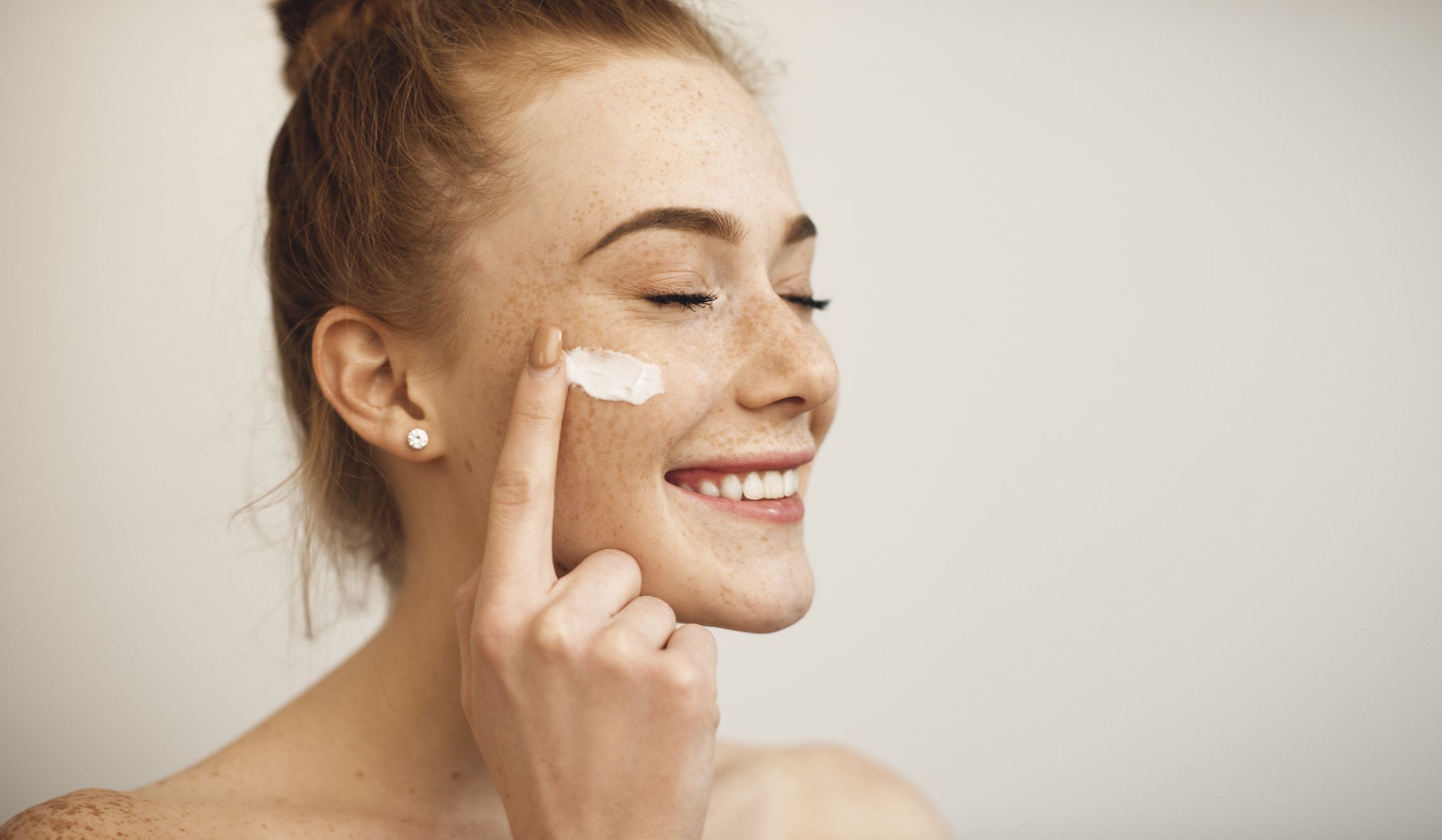 close up of a young female with red hair and freckles applying white cream on her face laughing with closed eyes isolated on white background